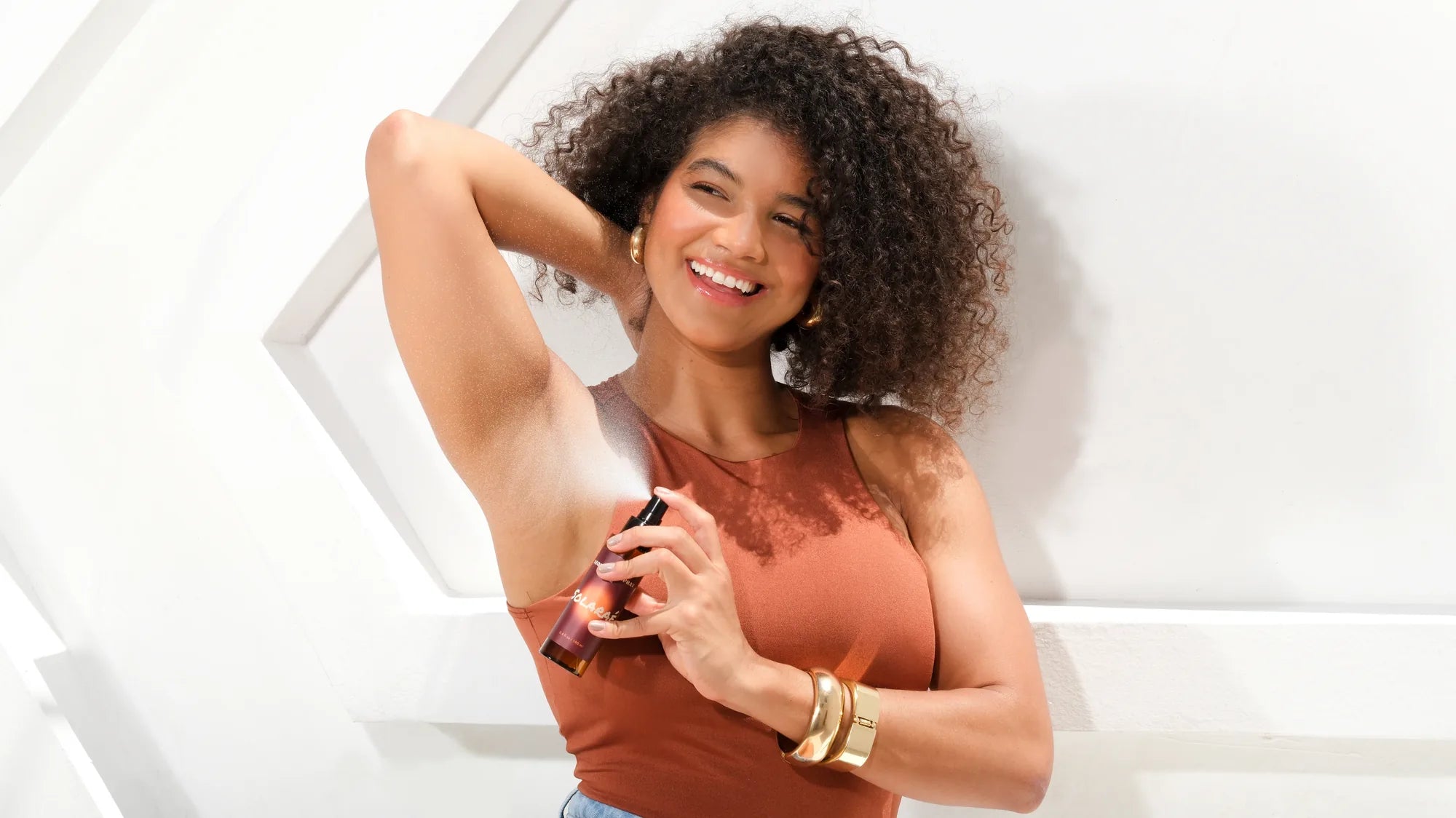Modelo de pele parda com cabelo cacheado aplicando o Solaraé Desodorante sem alumínio Oud na axila, fragrância amadeirada feminina